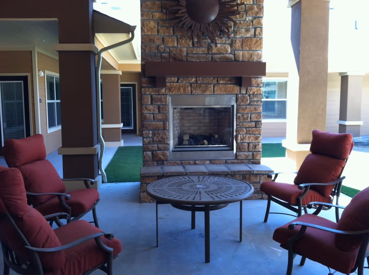 Outdoor seating area with four cushioned chairs arranged around a round metal table in front of a stone fireplace with a decorative sun wall hanging above it. The area is covered and surrounded by building walls and windows.