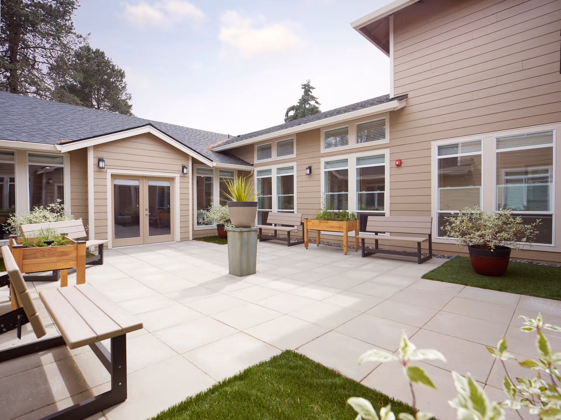 Outdoor courtyard area at Murray Highland Memory Care featuring beige building walls with multiple windows, several benches, potted plants, and a tiled floor under a partly cloudy sky.