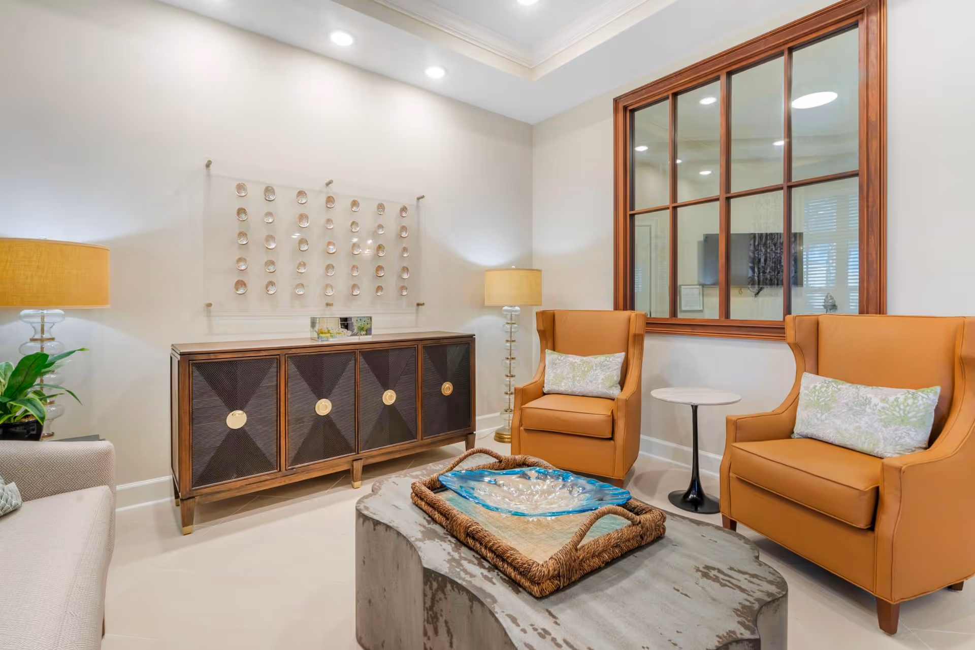 A bright living room seating area with two tan wingback chairs, a wooden sideboard, table lamps, and a central coffee table with a decorative tray.