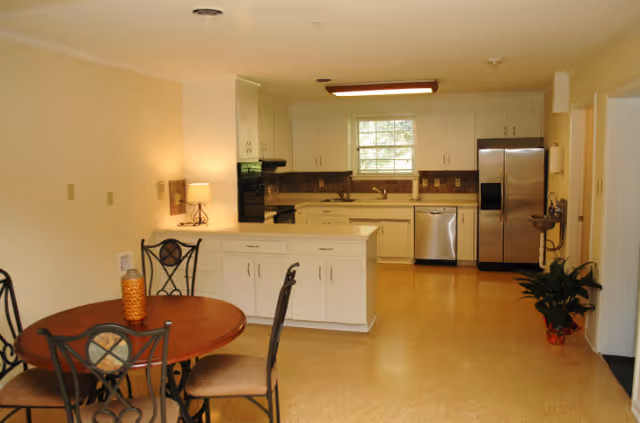 Open kitchen and dining area with a round wooden table and chairs in the foreground, white cabinets, a window above the sink, and stainless steel appliances.