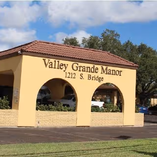 Exterior view of the entrance sign for Valley Grande Manor Nursing and Rehabilitation, displaying the address 1212 S. Bridge, with a tiled roof and two archways, surrounded by greenery and trees under a partly cloudy sky.