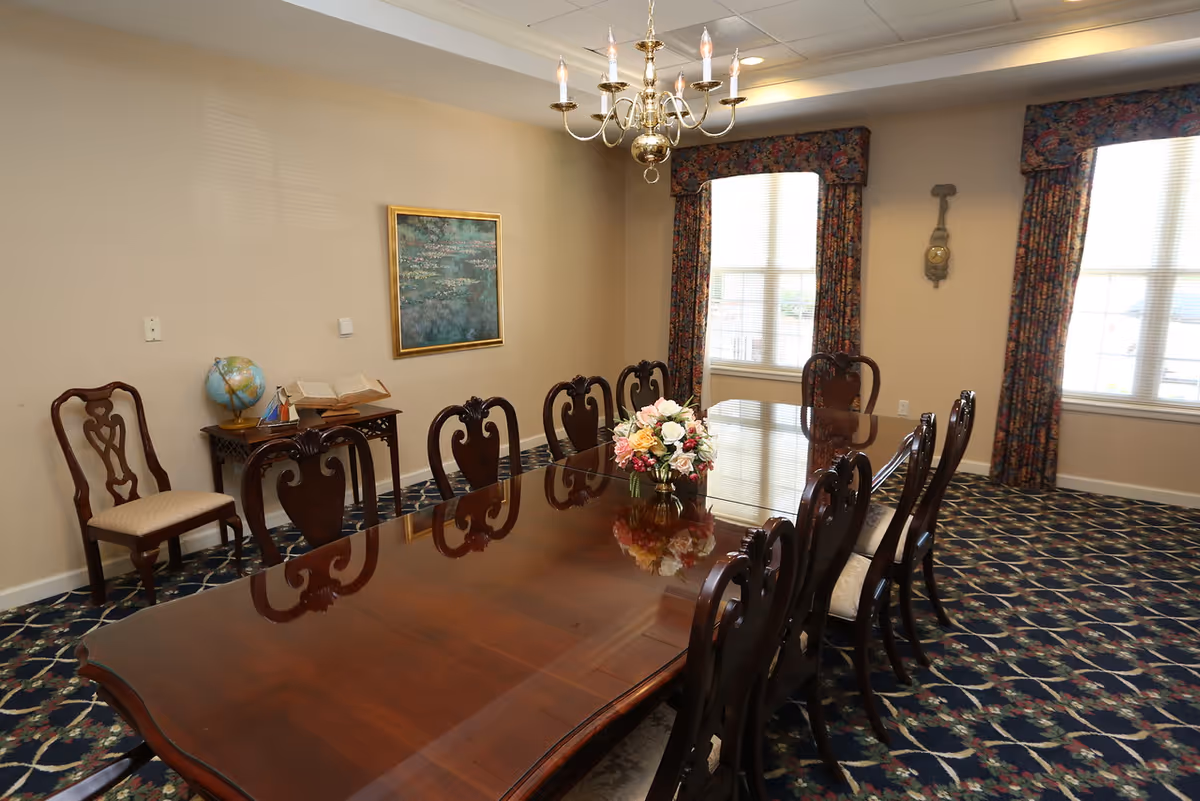 A formal dining room with a long polished wooden table surrounded by carved chairs, a floral centerpiece, chandelier, windows with patterned curtains, and a side table with a globe.
