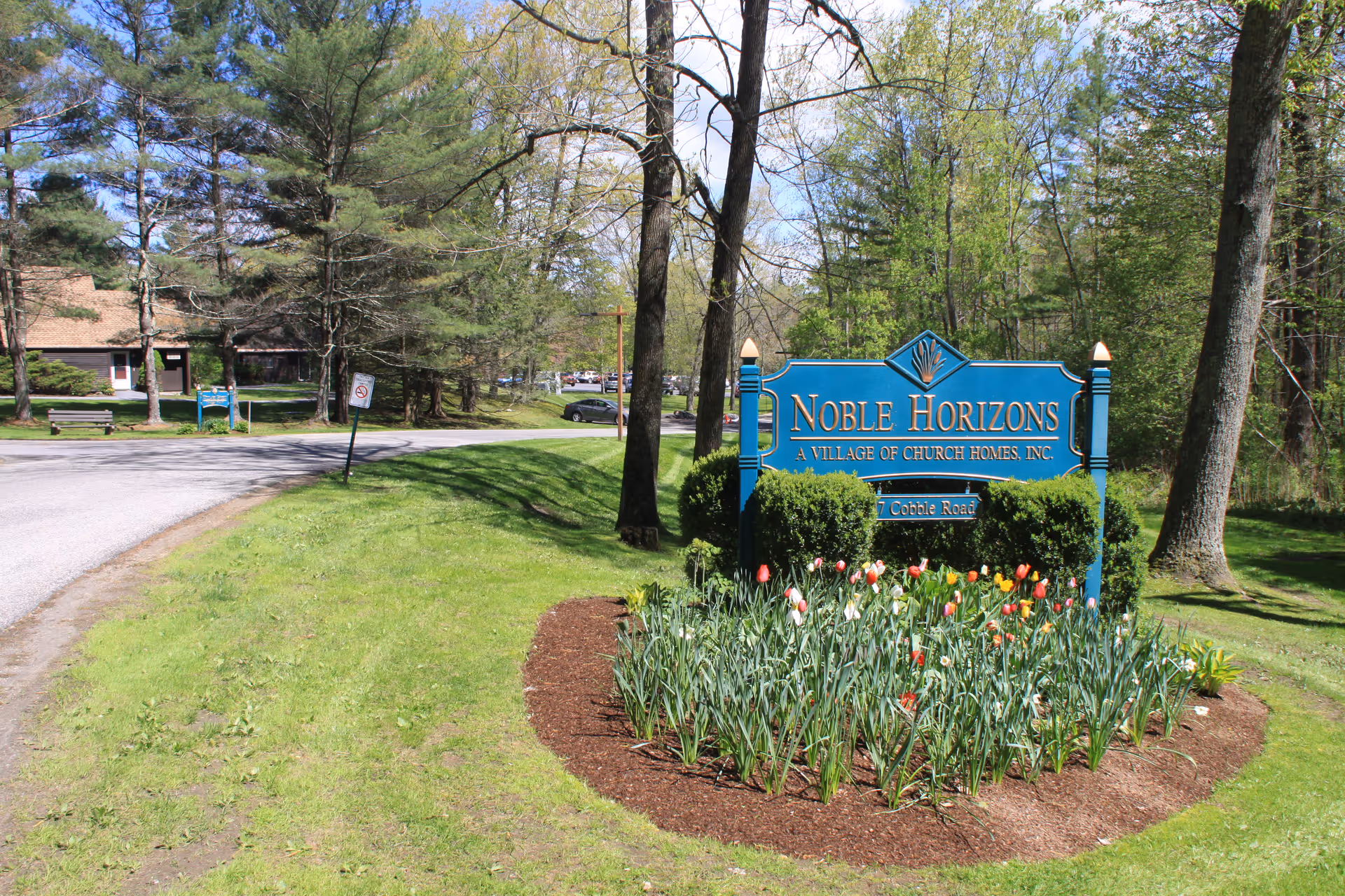 Blue 'Noble Horizons' entrance sign surrounded by a tulip flowerbed, lawn, trees, and a driveway with buildings in the background.