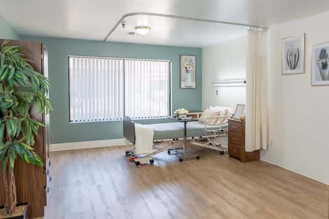 A bright, spacious senior care bedroom with a hospital-style bed, bedside table, privacy curtain, and a large window with vertical blinds.
