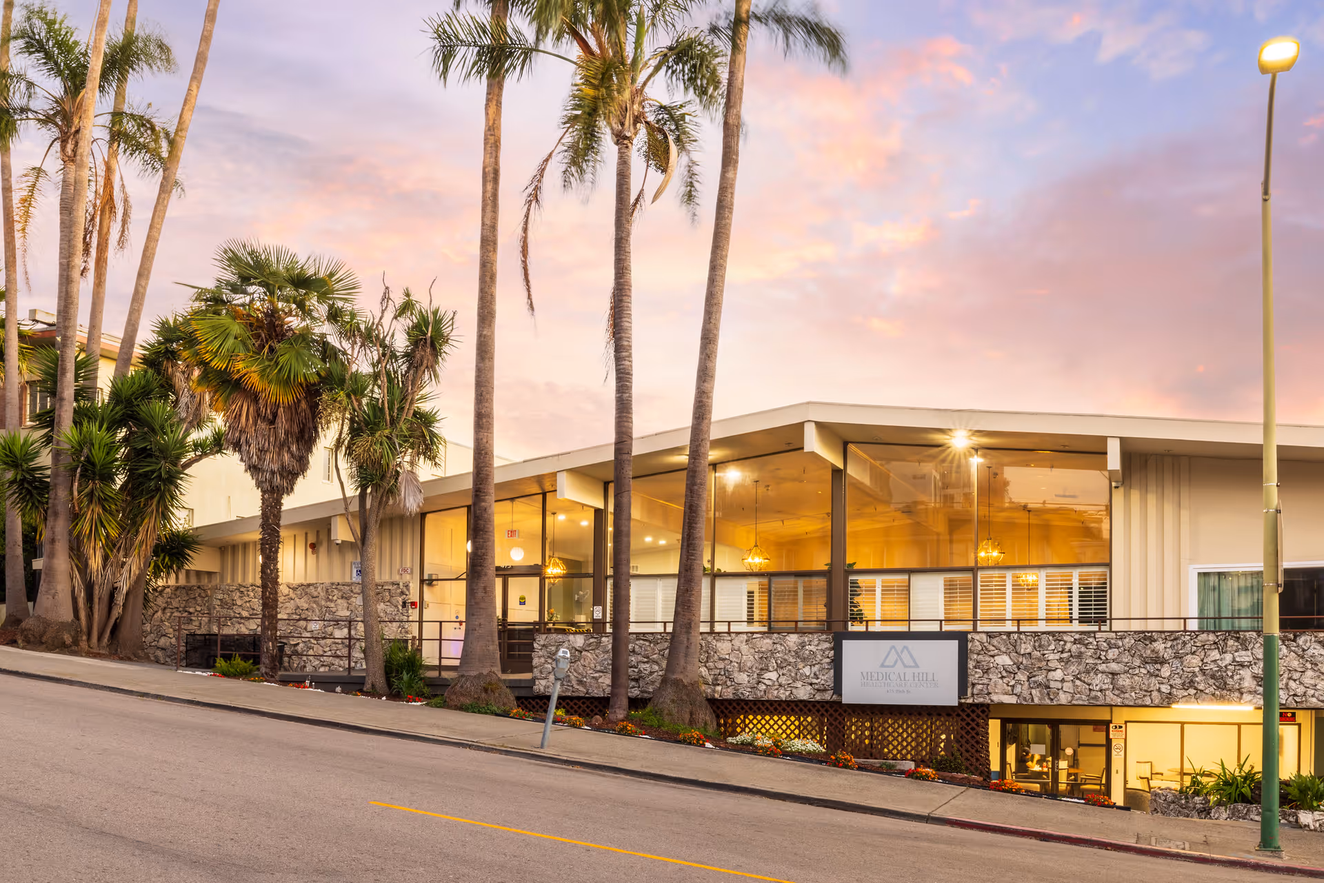 Front exterior of a mid-century modern healthcare building with large glass windows, stone facade and palm trees at sunset.