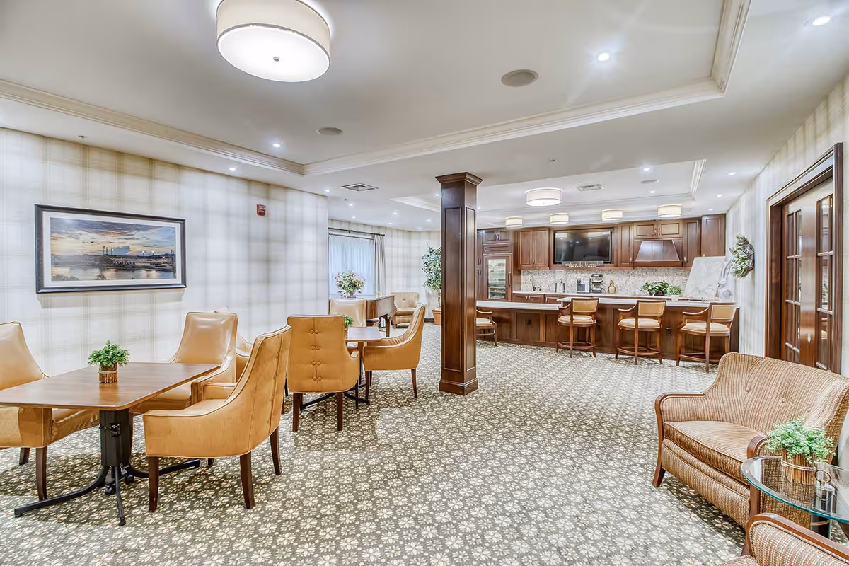 Bright communal dining and lounge area with tables, upholstered chairs, and a serving bar/kitchen.