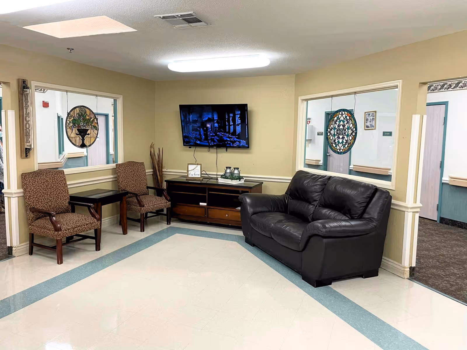 A nursing facility common area with a black leather couch, two patterned armchairs, a wooden TV stand with decorative items, and a wall-mounted flat screen TV. The walls are beige with two large windows looking into adjacent hallways. There is a 'WELCOME' sign on the left wall and decorative stained glass hanging in the windows.