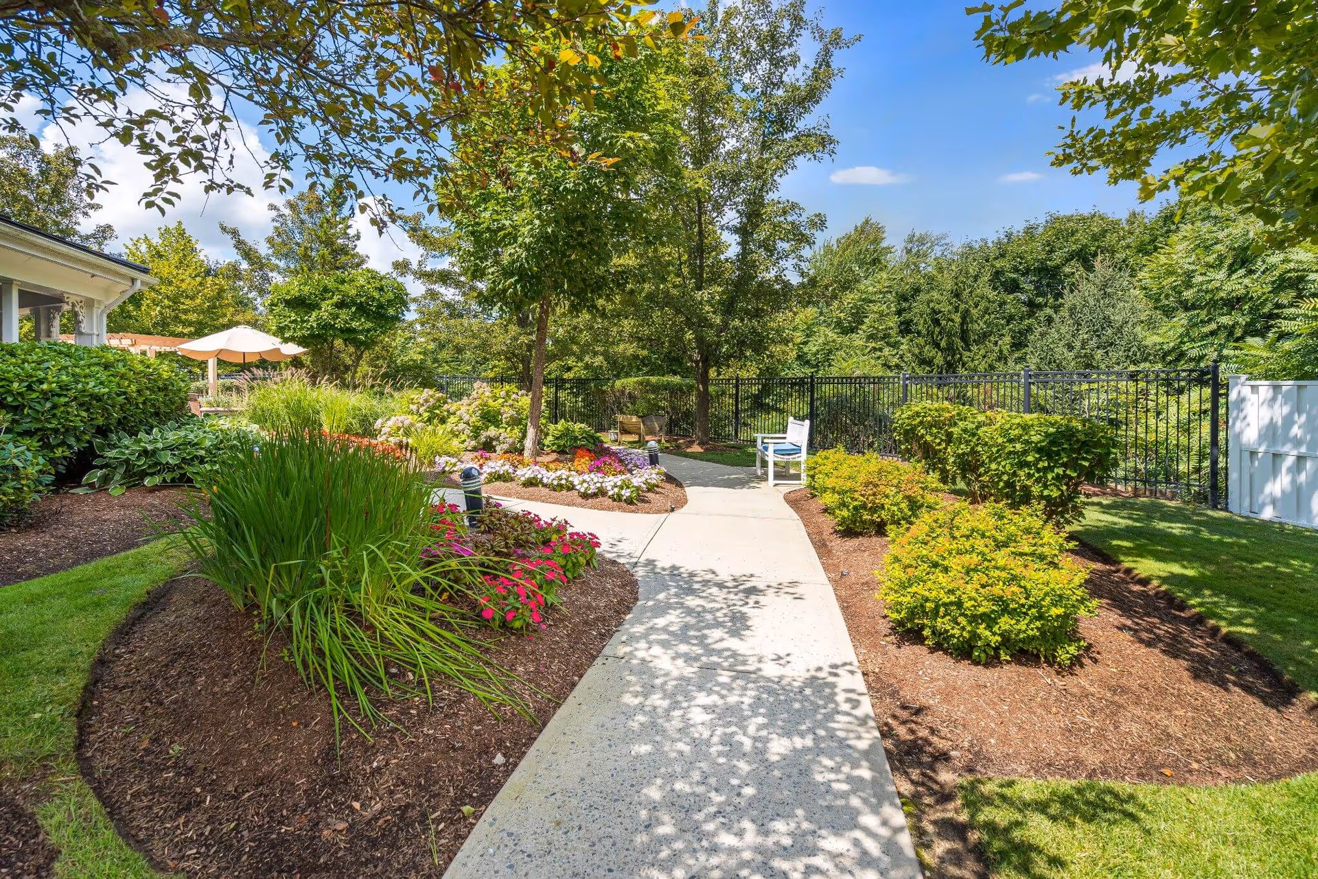 Sunlit landscaped garden path lined with flower beds, shrubs, a white chair and umbrella near a fence.