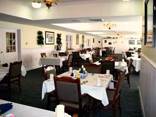 Dining room with multiple tables set with white tablecloths and chairs in a senior living facility.