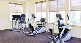 A bright exercise room with large windows covered by beige blinds. The room contains several pieces of cardio equipment including two elliptical machines and a recumbent bike, all positioned on a carpeted floor.
