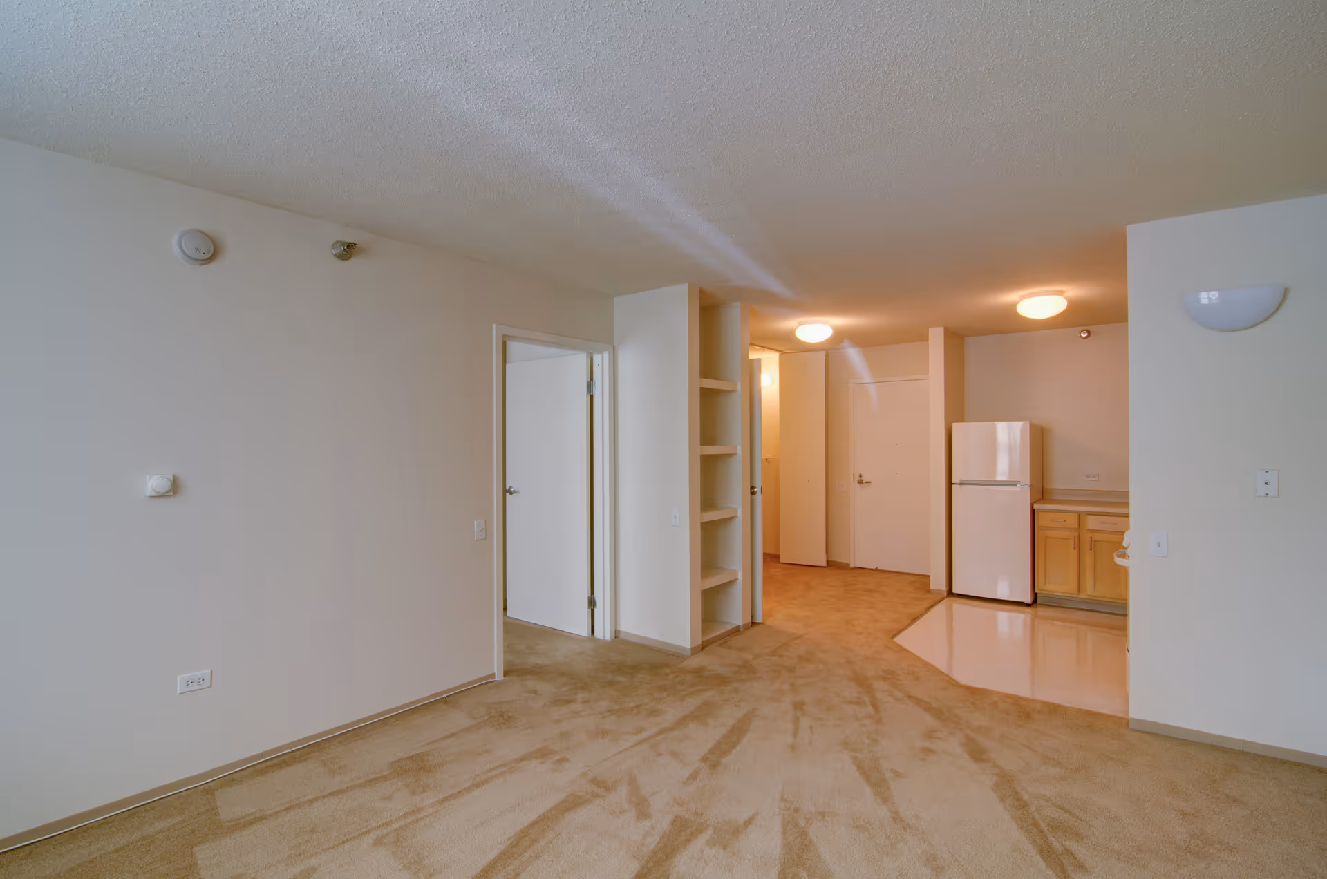Interior view of an unfurnished senior living apartment featuring beige carpeted flooring, white walls, a small kitchen area with a white refrigerator and wooden cabinets, open shelving, and a doorway leading to another room.