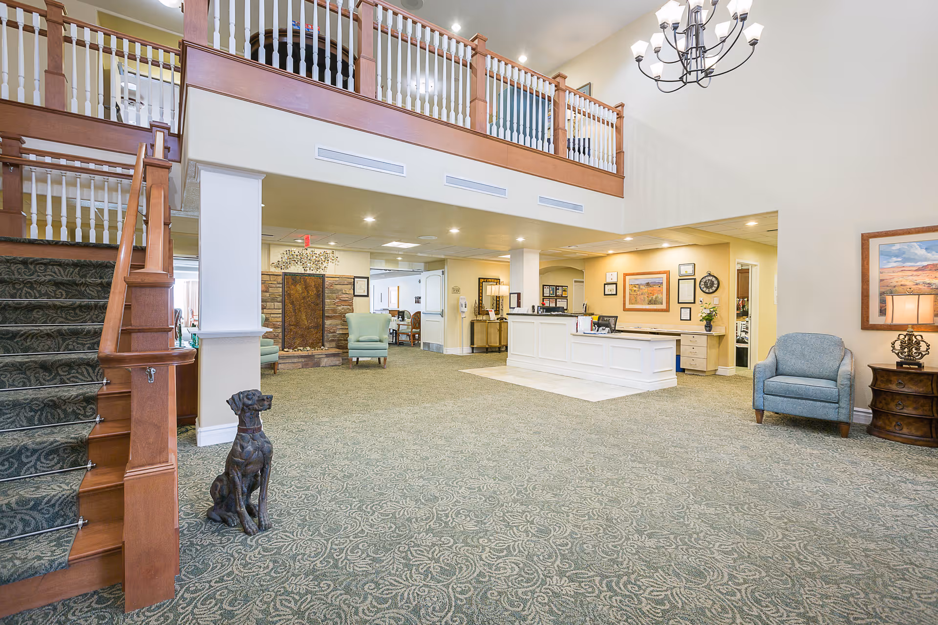 Spacious senior living facility lobby with a patterned carpet, wooden staircase with carpet runner, a bronze dog statue, a white reception desk, light blue armchairs, framed artwork on the walls, and a chandelier hanging from the high ceiling.