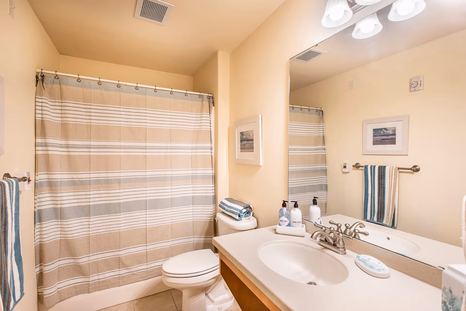 A clean bathroom with a beige and white striped shower curtain, a white toilet, a countertop with a sink, soap dispensers, and a folded towel. There is a large mirror above the sink reflecting the shower curtain and a framed picture on the wall. The walls are painted light beige.