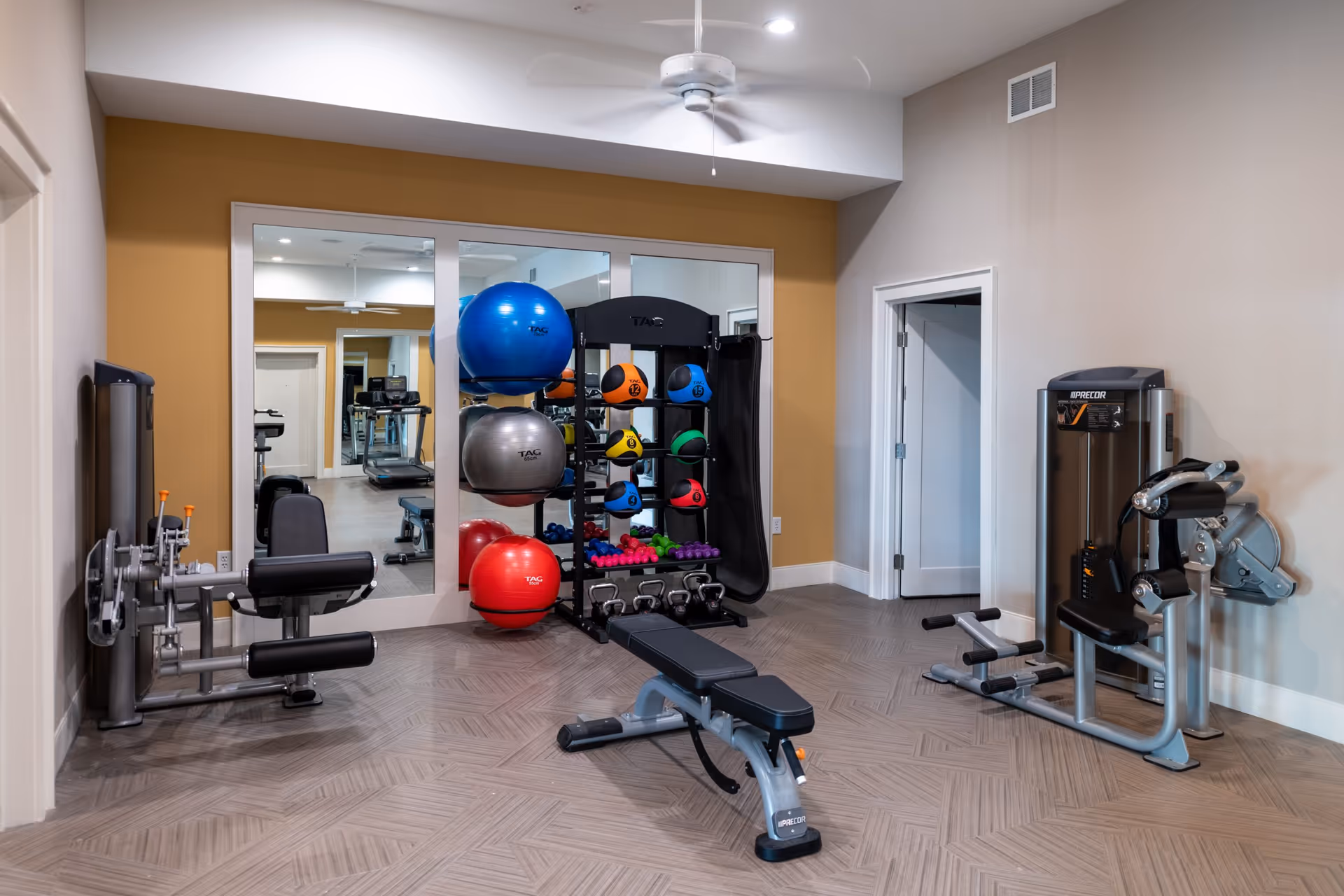 A fitness room with exercise equipment including a weight bench, leg curl machine, and a rack holding colorful exercise balls, medicine balls, dumbbells, and kettlebells. The room has a large mirror on one wall, a ceiling fan, and light-colored walls with a mustard accent wall.