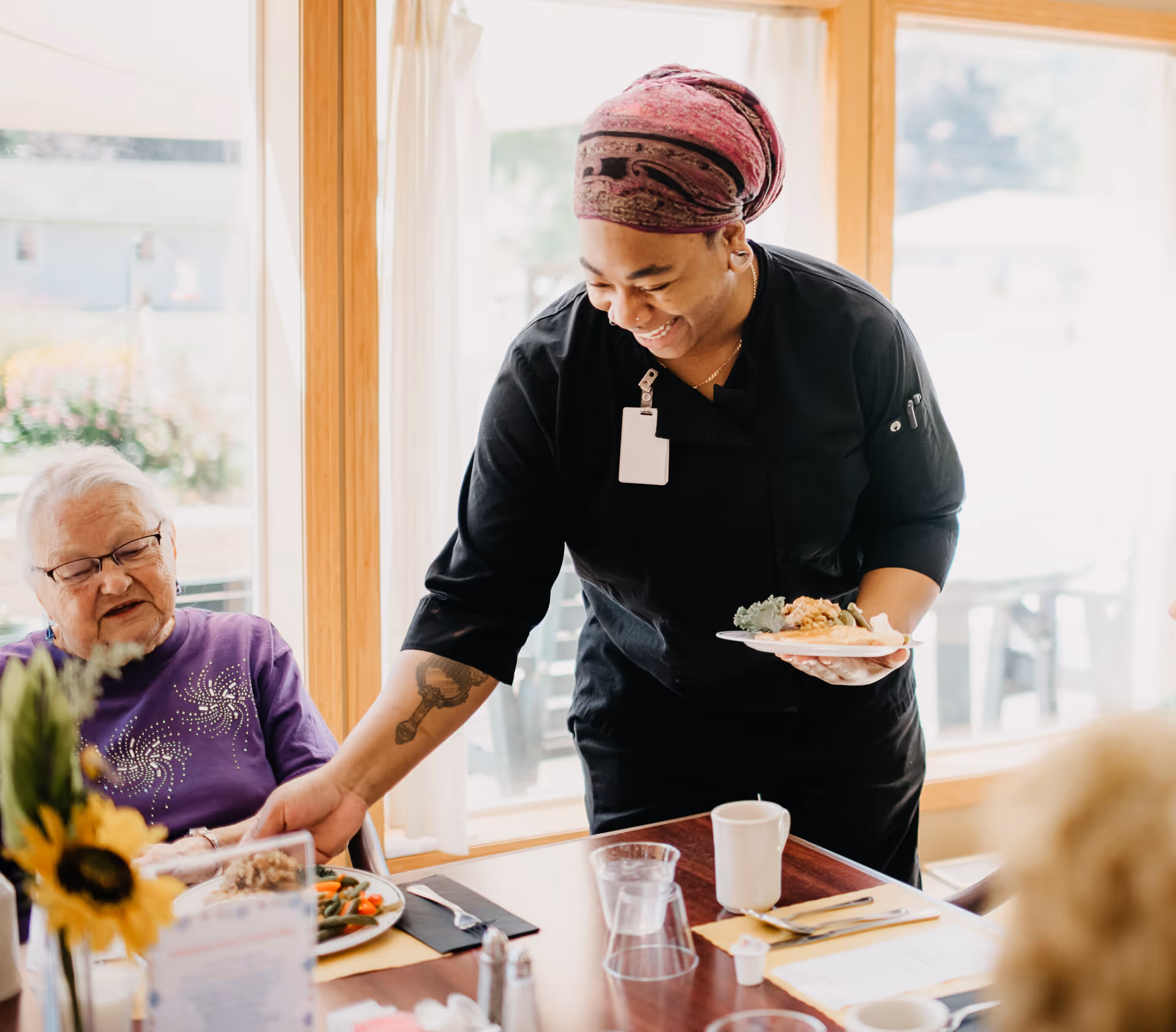 A smiling staff member wearing a black uniform and a purple headscarf serves a plate of food to an elderly woman seated at a dining table with plates of food, cups, and a sunflower centerpiece near a large window with white curtains.