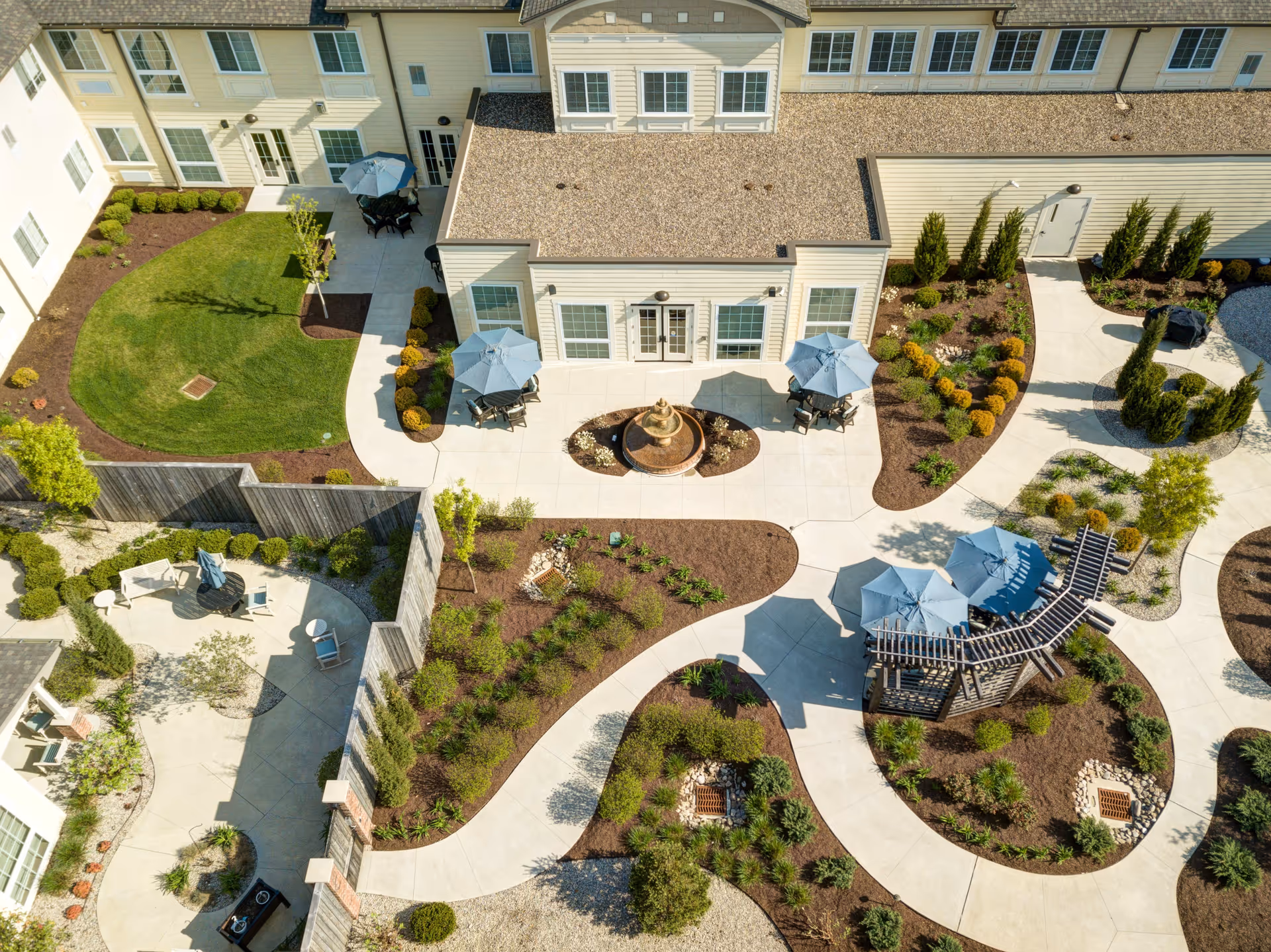 Aerial view of an outdoor courtyard at Traditions of West Chester featuring paved walkways, landscaped garden beds, green lawns, patio tables with umbrellas, a central fountain, and a wooden pergola structure.