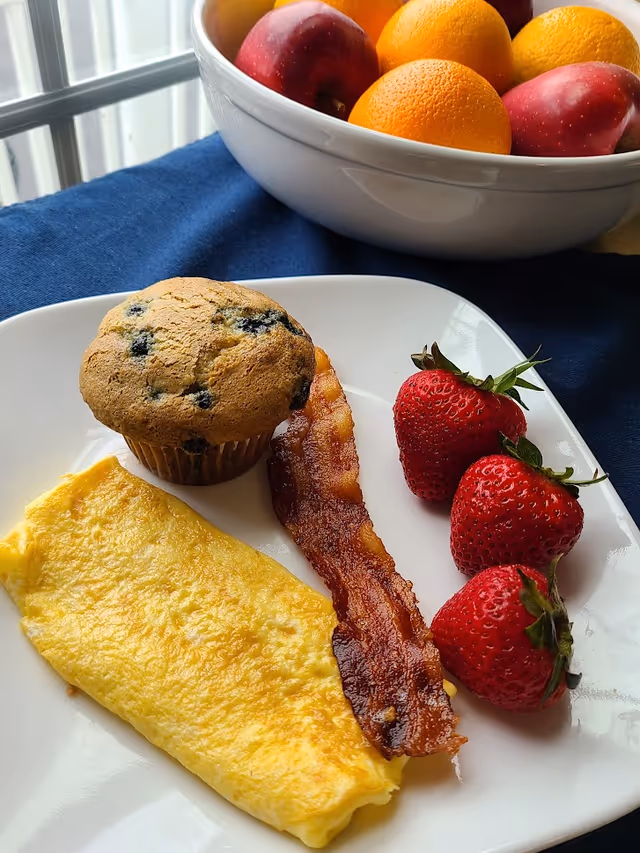 A white plate with a folded yellow omelette, a strip of crispy bacon, three fresh strawberries, and a blueberry muffin. In the background, there is a white bowl filled with red apples and oranges on a blue tablecloth near a window.
