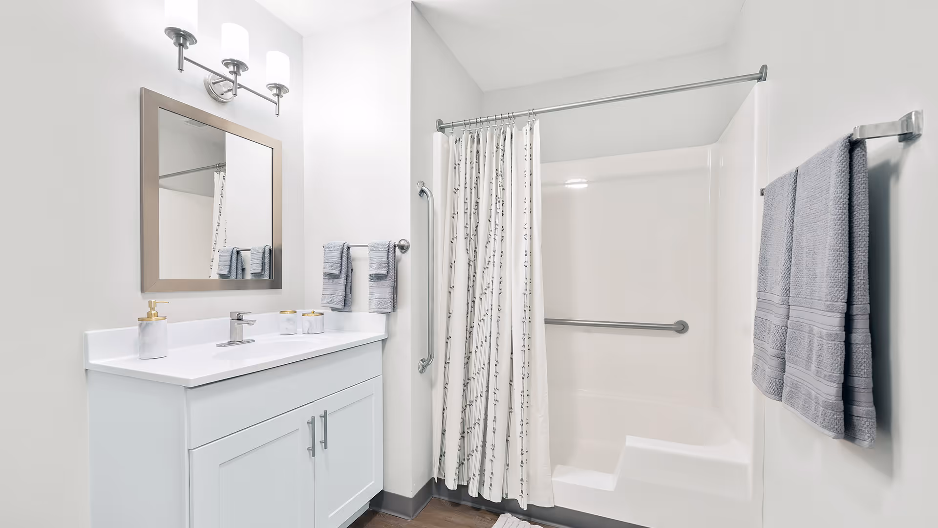 A clean and modern bathroom featuring a white vanity with a sink, a large mirror with a silver frame, and two wall-mounted light fixtures above. There are gray towels hanging on towel racks, a white shower curtain with a subtle pattern, and a built-in bathtub with grab bars for safety.