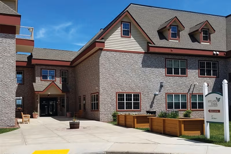 Exterior view of a senior living facility building with brick walls and multiple windows. The entrance is visible with a covered walkway, potted plants, and a bench nearby. A sign in the foreground reads 'Hyland Park' with a welcome message. The sky is clear and blue.