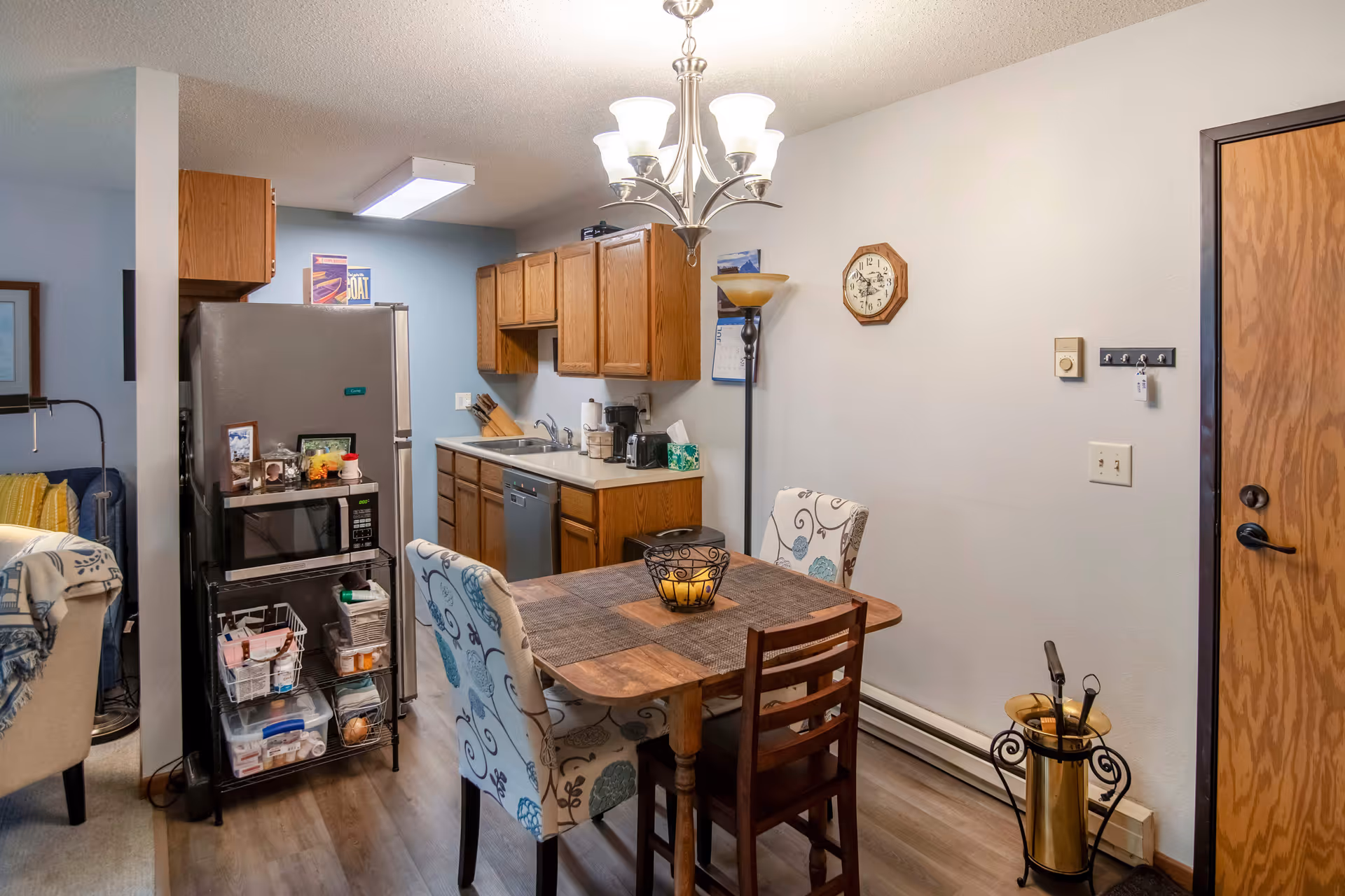 A small kitchen and dining area in a senior living facility. The kitchen has wooden cabinets, a stainless steel refrigerator, a microwave on a metal rack, a dishwasher, and a sink. The dining area features a wooden table with four chairs, two of which have floral upholstery. A floor lamp and a wall clock are visible on the right wall, along with a wooden door. The flooring is wood-style laminate.
