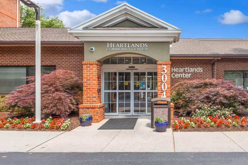 Front entrance of Heartlands Community Center at 3004, with brick walls, glass double doors, landscaped flower beds with red and white flowers, and two potted plants on either side of the entrance under a clear blue sky.