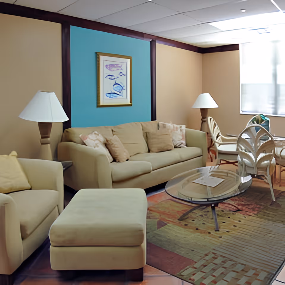 A cozy living room with beige sofas, an ottoman, glass coffee table, lamps, and chairs by a window.