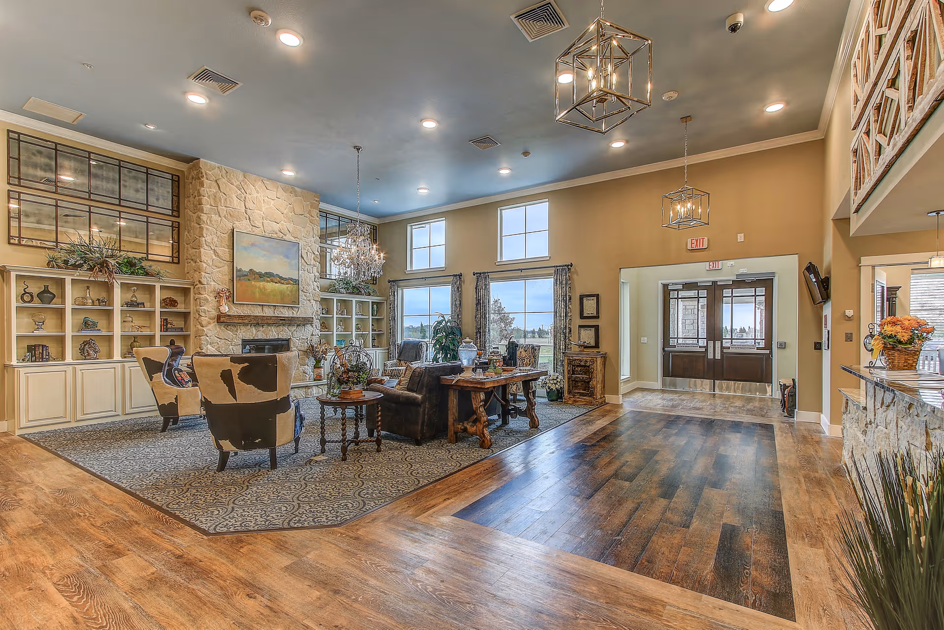 Spacious and well-lit living room area with wooden flooring, a stone fireplace, and large windows letting in natural light. The room features comfortable seating including armchairs and a sofa arranged around a coffee table, decorative shelves with various ornaments, and elegant chandeliers hanging from the ceiling. The entrance door is visible in the background.