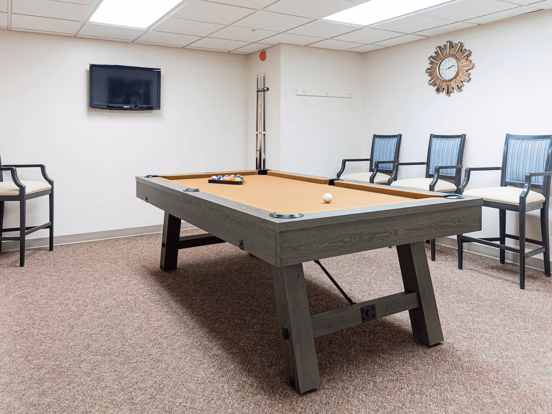 A game room with a pool table in the center, a wall-mounted TV, several chairs along the wall, a wall clock, and pool cues standing in the corner.