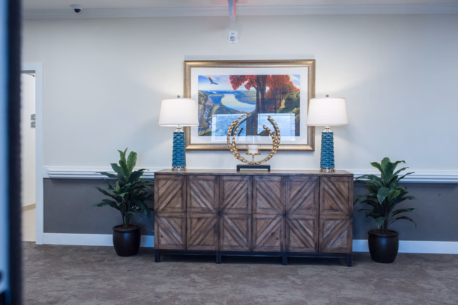 Wooden console table with two lamps, decorative sculpture, framed artwork and potted plants in a well-lit interior hallway.