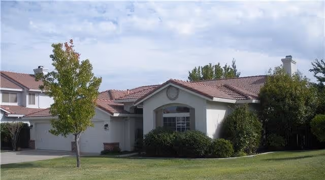 Single-story residential building with a tiled roof, surrounded by green grass, bushes, and a tree with some autumn-colored leaves under a partly cloudy sky.
