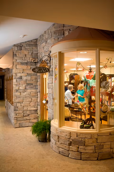 Interior view of a market area inside a senior living facility with stone walls and a large window display featuring clothing and hats. Two elderly women are seen inside the market talking to each other.