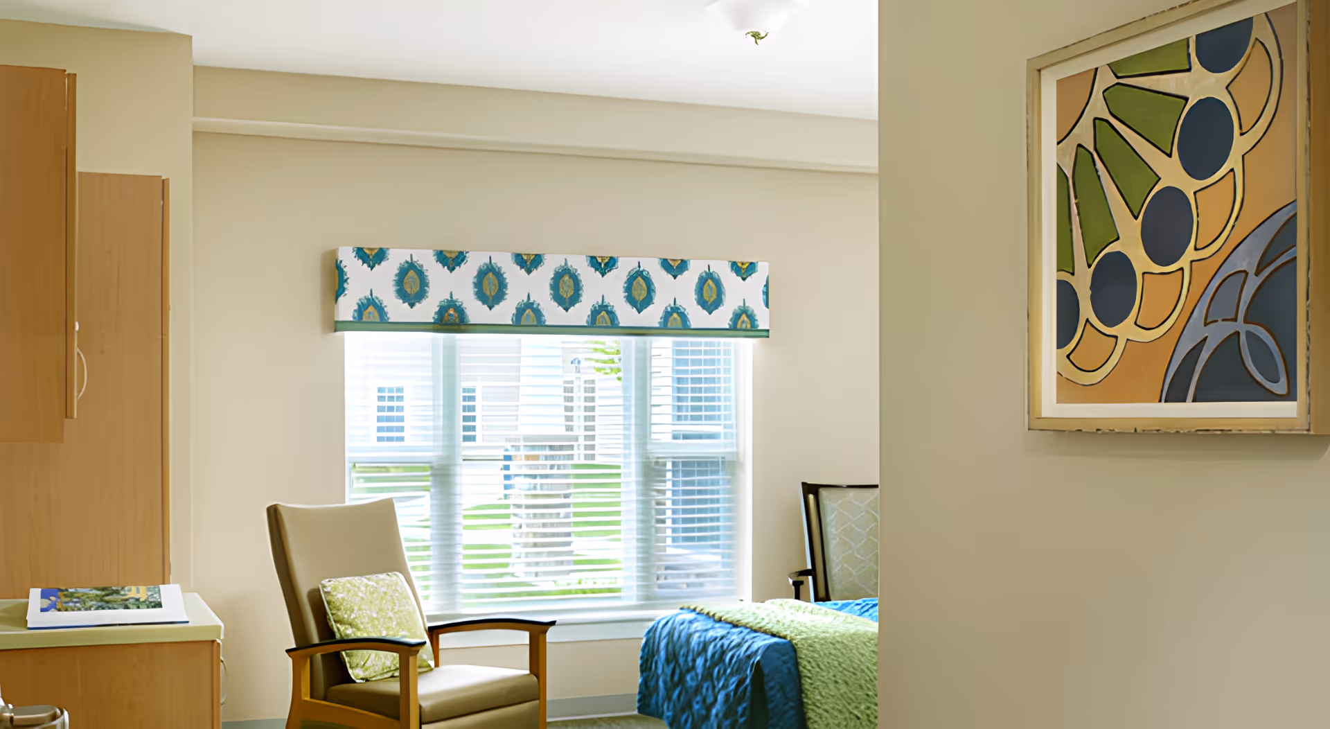 Sunlit bedroom with a chair, window with a patterned valance, and a partially visible bed and bedside furniture.