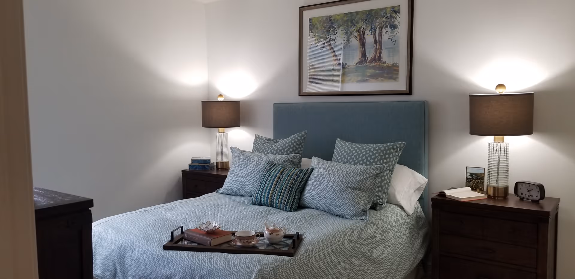 A neatly made bed with a light blue upholstered headboard and multiple decorative pillows in shades of blue and white. There are two dark wooden nightstands on either side of the bed, each with a lamp featuring a clear base and a dark lampshade. On the left nightstand, there are two stacked books, and on the right nightstand, there is an open book, a small framed picture, and a clock. Above the bed hangs a framed painting of trees and a landscape. The room has white walls and soft lighting.