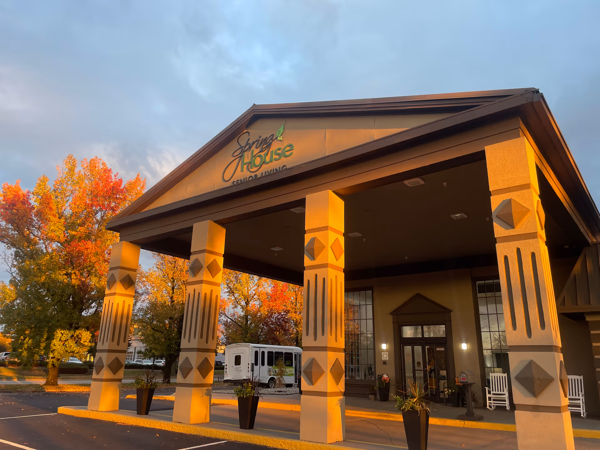 Front entrance of The Spring House senior living building with a covered porte-cochere and decorative columns lit by warm sunset light.