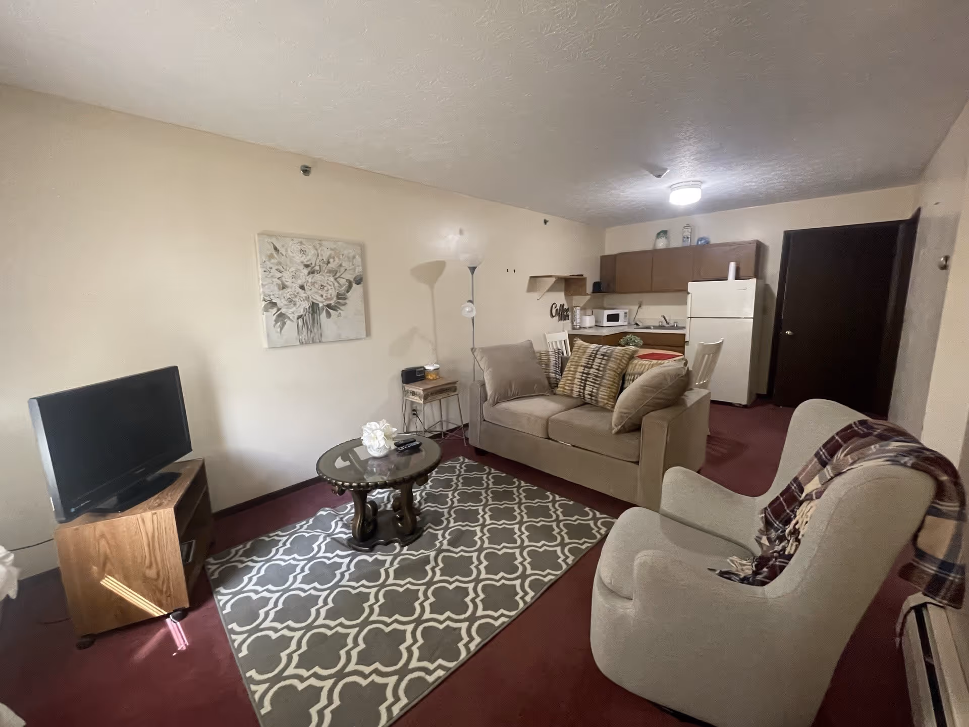 A cozy living area in an assisted living facility featuring a beige loveseat with patterned pillows, a beige armchair with a plaid throw draped over it, a round glass coffee table on a patterned rug, a wooden TV stand with a flat-screen TV, and a small kitchen area in the background with a refrigerator, microwave, sink, and cabinets.