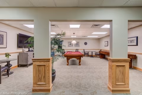 Interior view of a recreational room in a senior living facility with a pool table in the center, a piano against the far wall, and several chairs and tables around the room. The room has beige walls, carpeted floor, and ceiling lights. There are wooden columns and trim, a potted plant, and a television visible in an adjacent room.