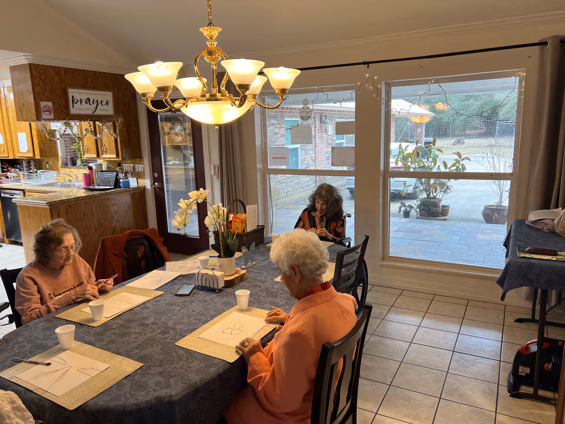 Three elderly women seated around a dining table doing activities under a chandelier with large windows looking outside.