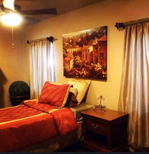 A cozy bedroom with a single bed dressed in red bedding and pillows. The room features two windows with light-colored curtains, a ceiling fan with a light, a wooden nightstand with a decorative metal cross, a potted green plant, and a large painting of a scenic village on the wall above the bed.