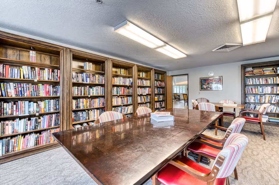 A bright room with large wooden bookshelves filled with books lining the walls. In the center is a large polished wooden table surrounded by chairs with striped upholstery. There is a smaller table with chairs in the background near a doorway leading to another room. The ceiling has fluorescent lighting and a clock and framed picture are visible on the far wall.