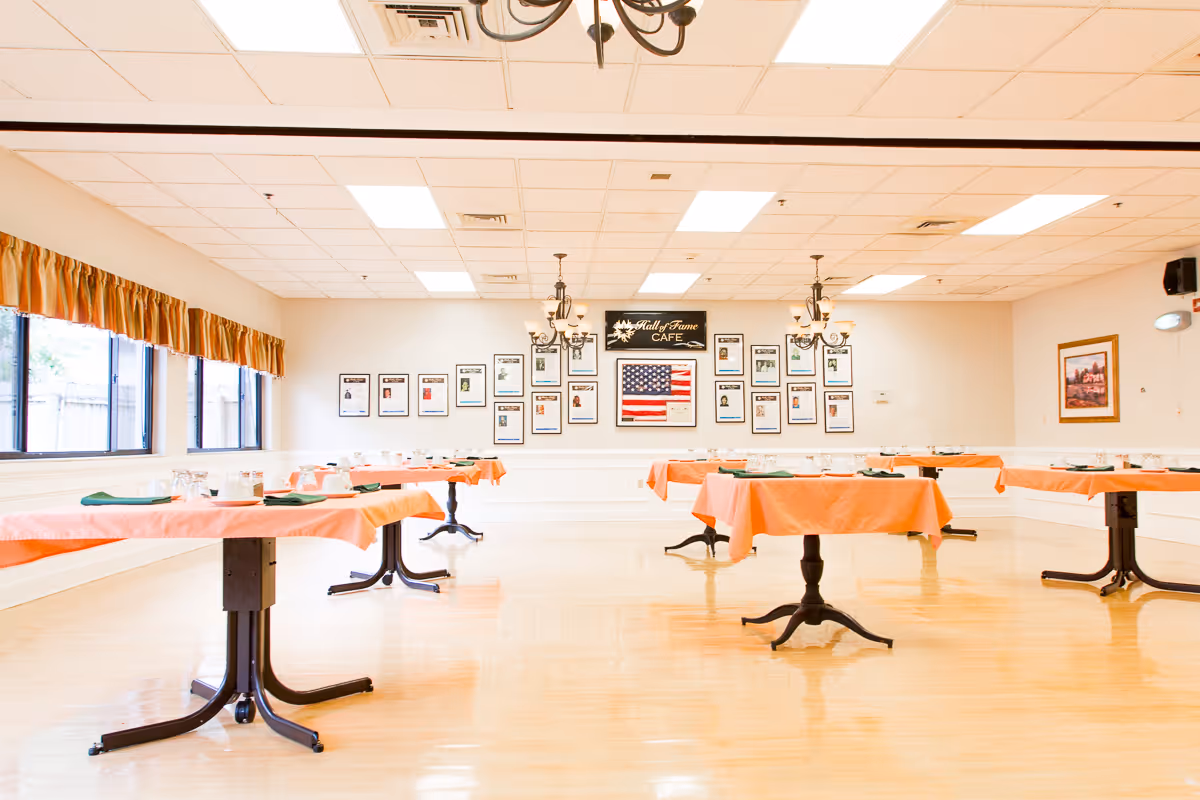 A bright dining room with several tables covered in orange tablecloths, each set with plates, cups, and green napkins. The room has large windows with orange curtains on the left side and a wall at the far end displaying framed photos and a sign that reads 'Hall of Fame Cafe' above an American flag. The floor is polished wood, and chandeliers hang from the ceiling.