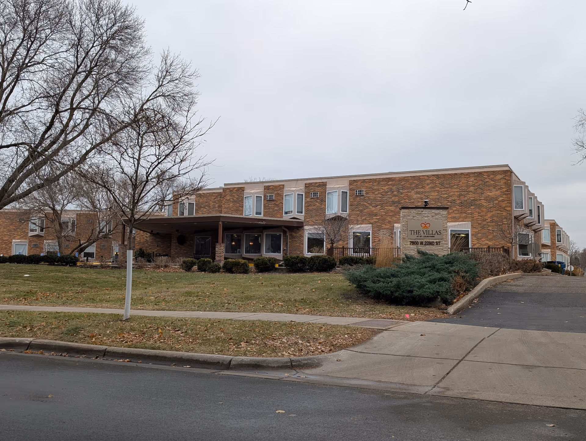 Two‑story brick senior living building with an entrance canopy, lawn and bare trees, and a sign reading 'The Villas' near the front.