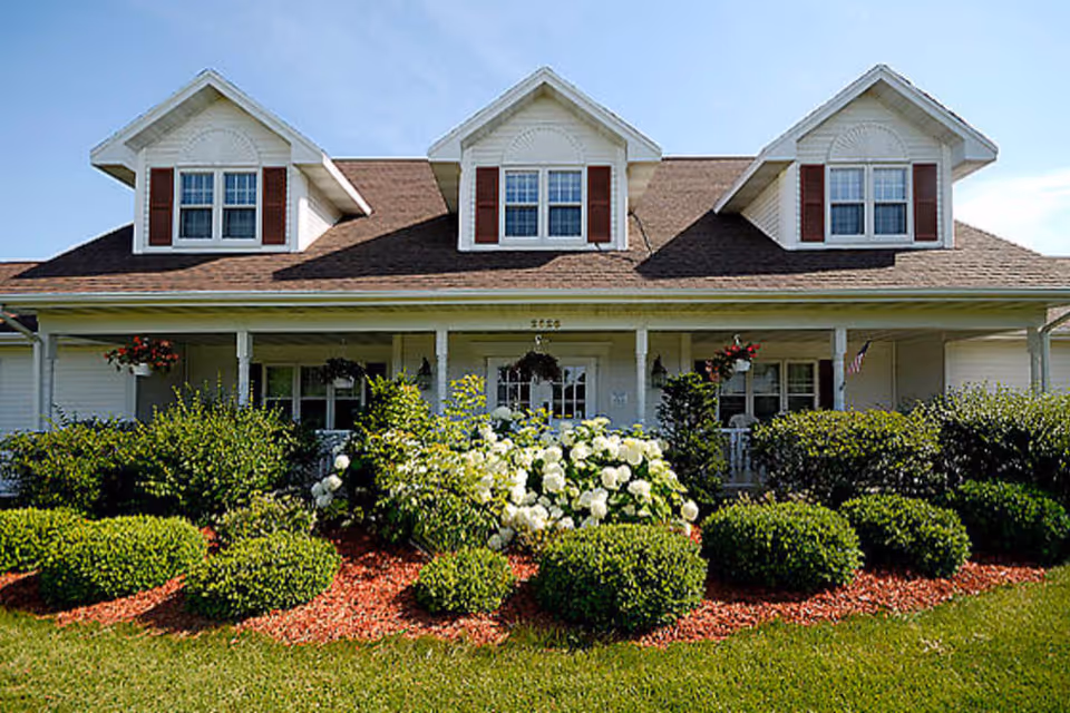 Front exterior of a two-story house-style senior living building with dormer windows, a covered porch and landscaped flower beds.