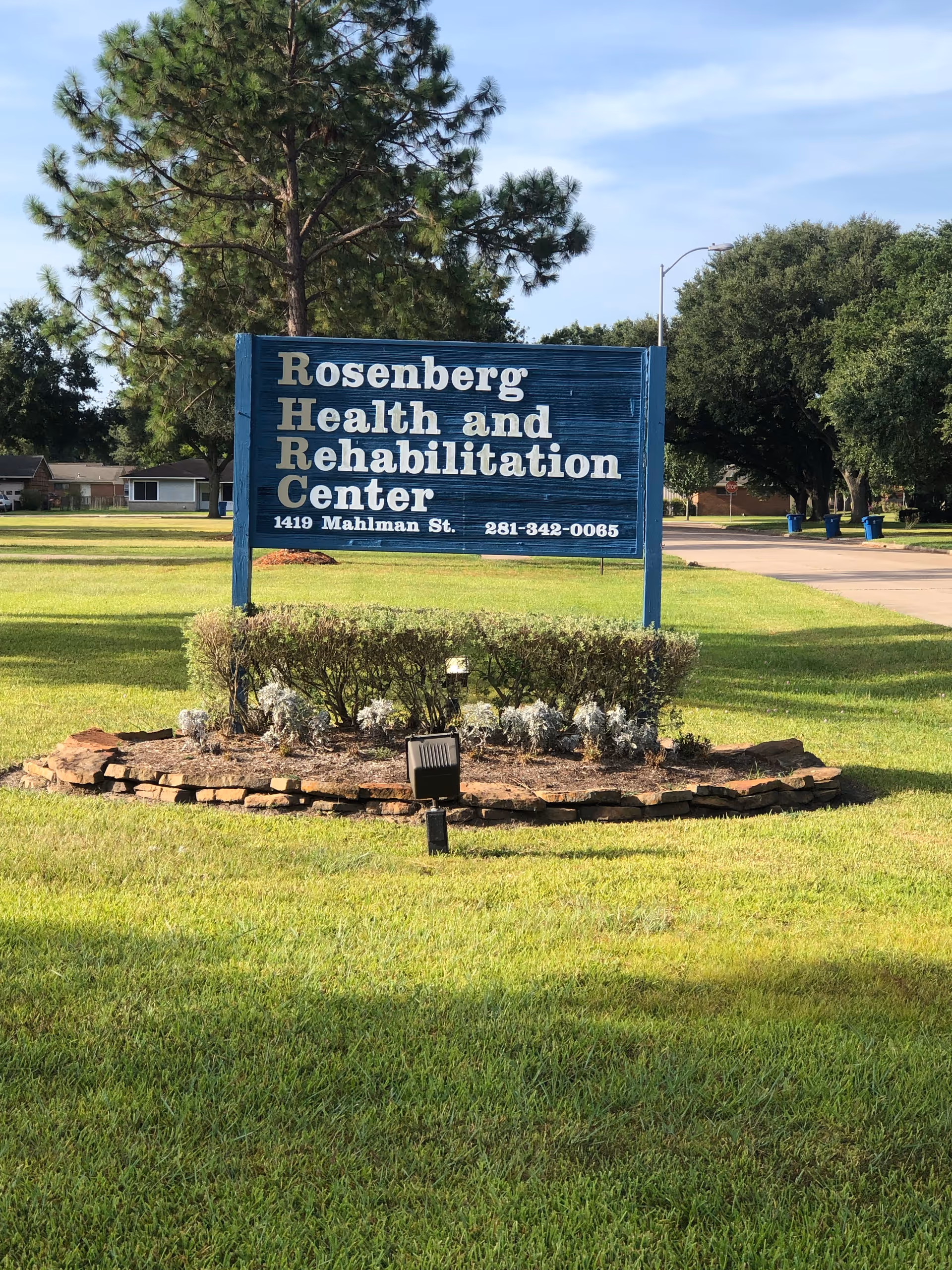 A blue wooden sign on a grassy lawn with bushes and a spotlight in front. The sign reads 'Rosenberg Health and Rehabilitation Center, 1419 Mahlman St., 281-342-0065'. Trees and a street are visible in the background under a partly cloudy sky.