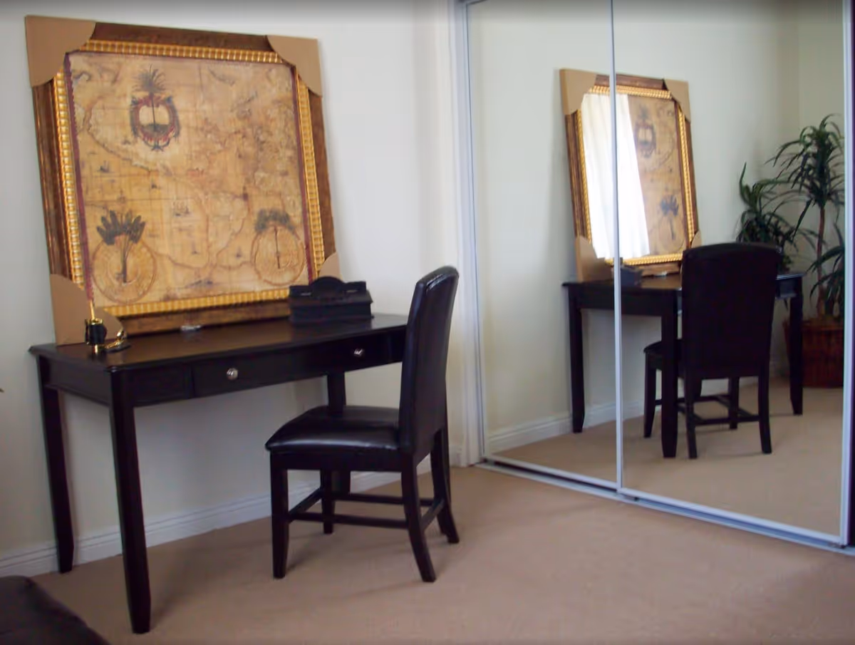 A room corner featuring a black wooden desk with two drawers, a black leather chair, and a large framed vintage map resting on the desk. The room has beige carpet flooring, white walls, a mirrored sliding closet door reflecting the desk and chair, and a potted plant in the corner.