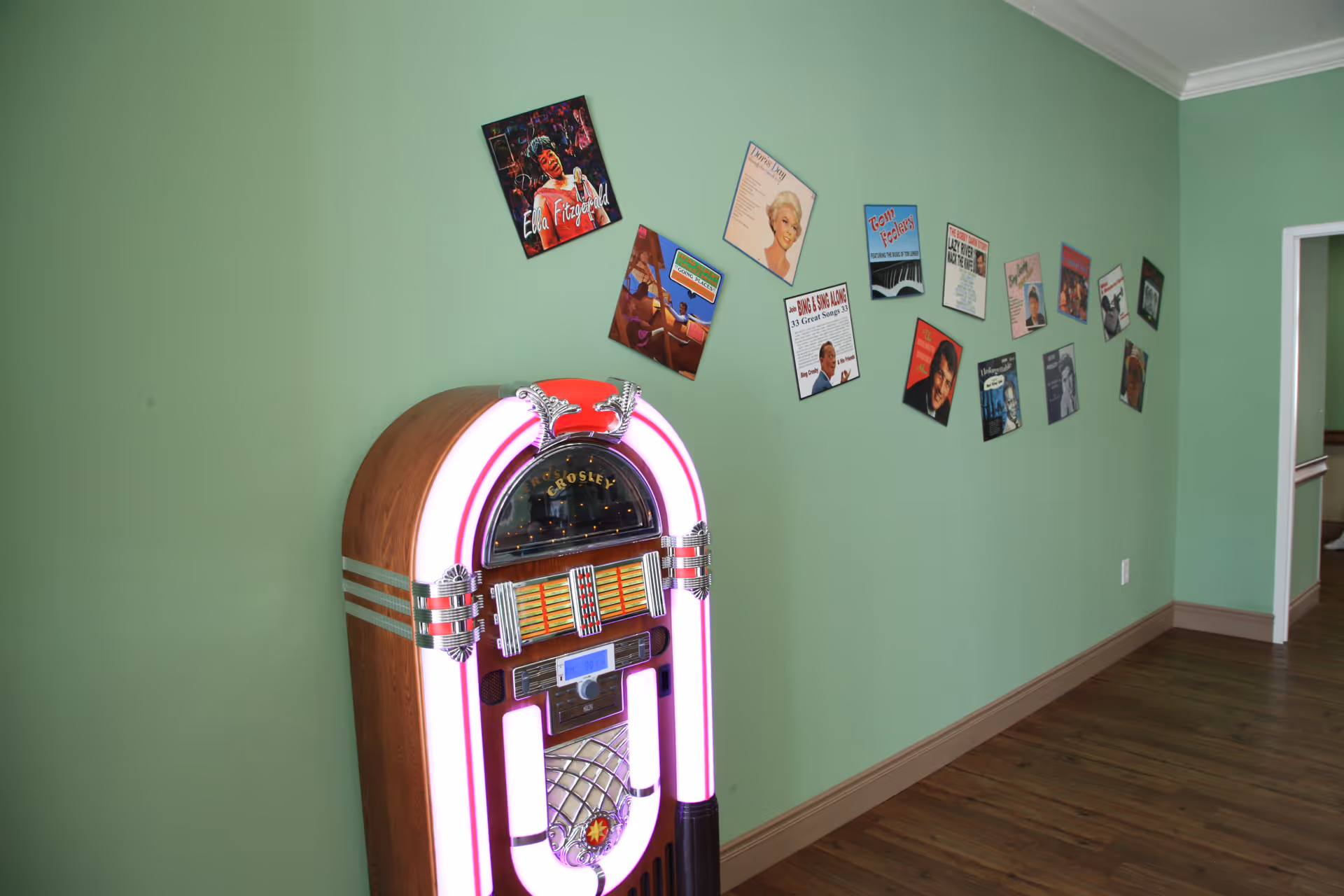 A vintage Crosley jukebox with pink neon lights stands against a green wall decorated with various retro music album covers. The floor is wooden, and there is an open doorway visible on the right side.
