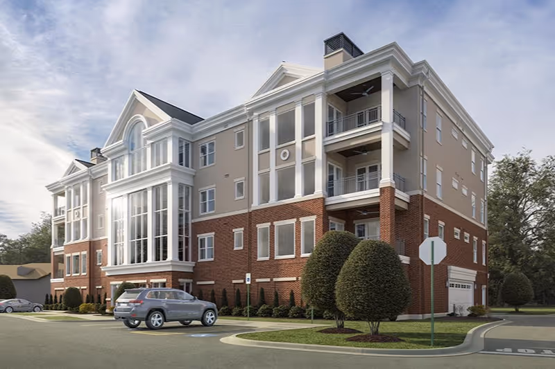 Exterior view of a multi-story senior living facility building with a combination of brick and beige siding, large windows, balconies, neatly trimmed bushes, and a parking area with cars.