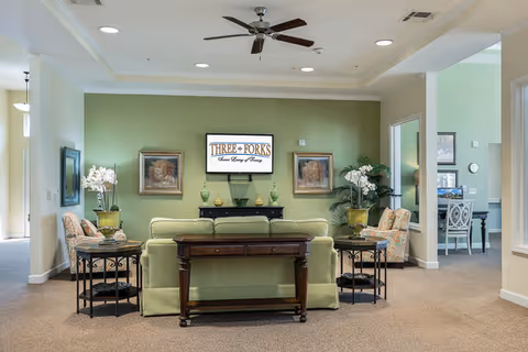 A cozy living room area with a green sofa facing a wall-mounted TV displaying the text 'THREE FORKS Senior Living & Memory'. The room features two patterned armchairs, side tables with decorative plants and flowers, framed artwork on the walls, a ceiling fan, and beige carpeting. There are open doorways leading to other rooms with light green walls.