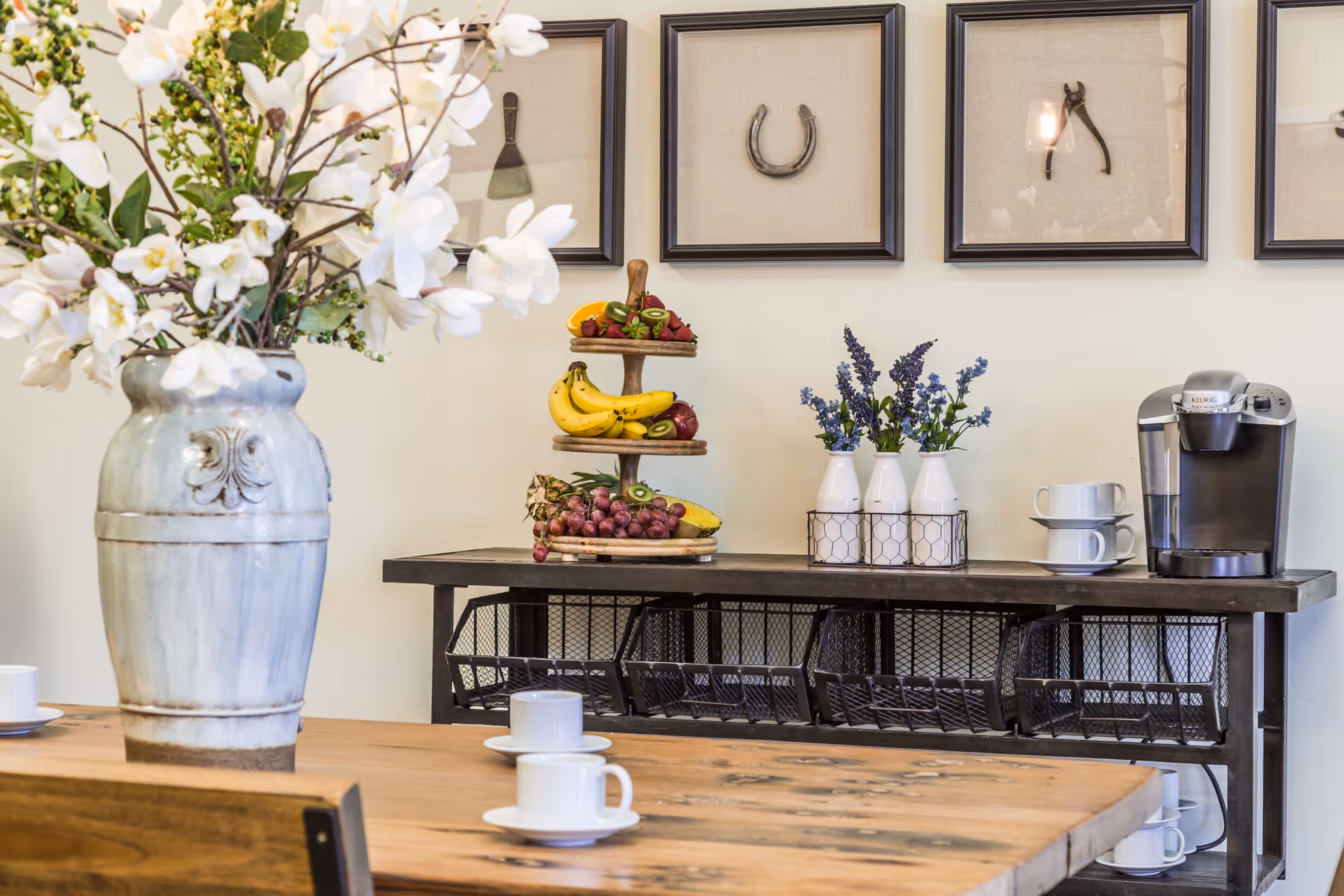 A communal dining area with a wooden table set with cups, a sideboard holding a coffee maker, fruit display, vases of flowers, and framed wall art.