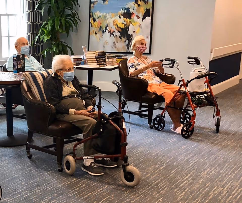 Three elderly individuals wearing face masks are seated in a common area of an assisted living facility. Two women are sitting in armchairs with walkers in front of them, and a man is seated at a table in the background. There is a table with a stack of books and a colorful abstract painting on the wall behind them.