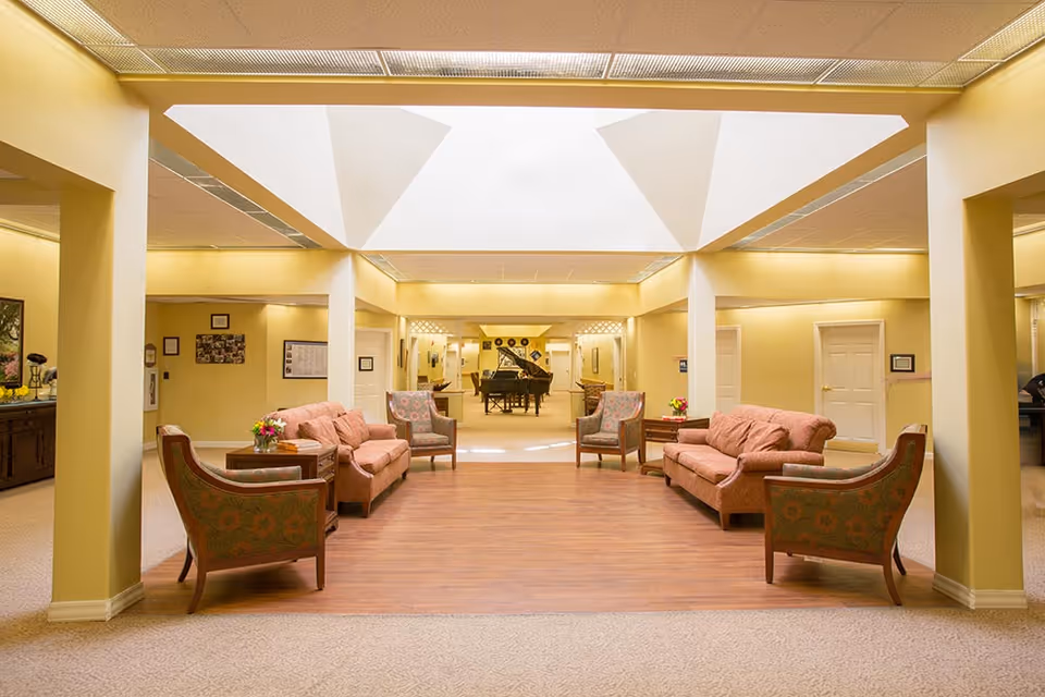 A spacious, well-lit common area with a skylight ceiling. The room features two pink sofas, four patterned armchairs, and small tables with flower arrangements. In the background, there is a grand piano and additional seating. The walls are painted light yellow, and the floor is a combination of carpet and wood.